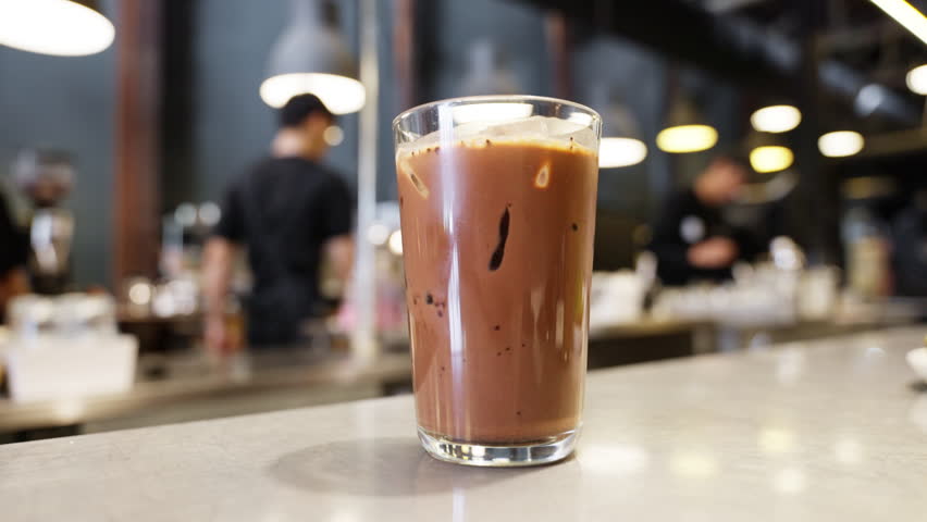 Indulgence awaits in a cafe setting with a tempting iced chocolate placed. The cool drink beckons against a backdrop of a bustling cafe, where a barista walks, adding an energetic touch to the scene. | Shutterstock HD Video #1111998417