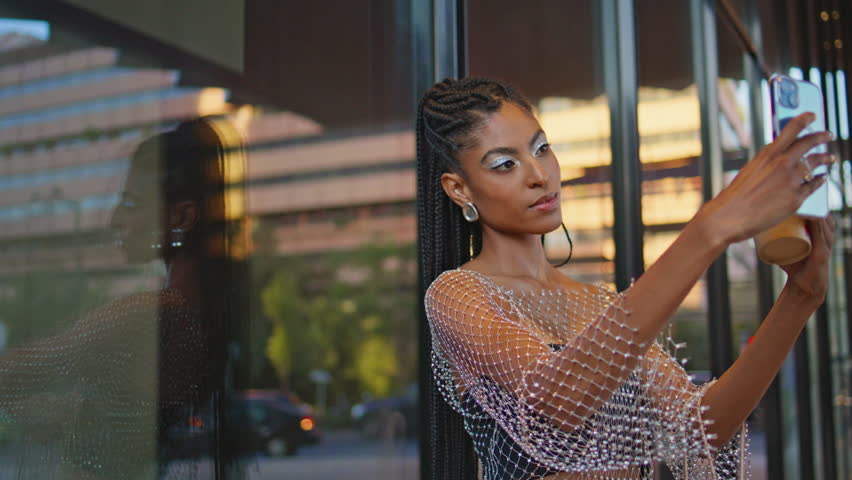 Pretty happy woman making selfie on glass wall view closeup. Stylish dark skin girl using smartphone taking picture herself on summer weekend. Positive smiling brunette posing with takeaway coffee
