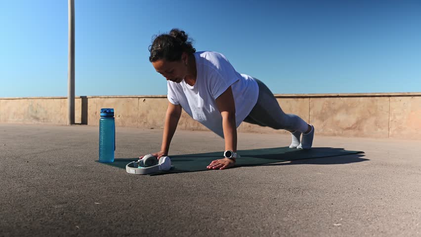 Multiracial middle aged active woman exercising outdoors, doing physical exercises on upper body, doing push ups on a fitness mat. Female athlete having difficulties performing bodyweight training | Shutterstock HD Video #1112001735