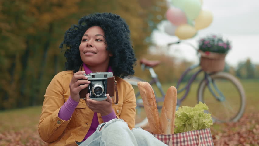 Smiling black lady with vintage camera capturing special moments of beautiful landscape in autumn park, sitting on grass with yellow fallen leaves. High quality 4k footage | Shutterstock HD Video #1112003245