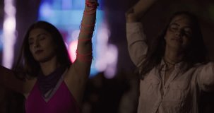 Two female friends dancing together on open space party with flashing lights. Happy beautiful girls with led rave lights on their hands. Out of focus crowd and big stage in the background.
