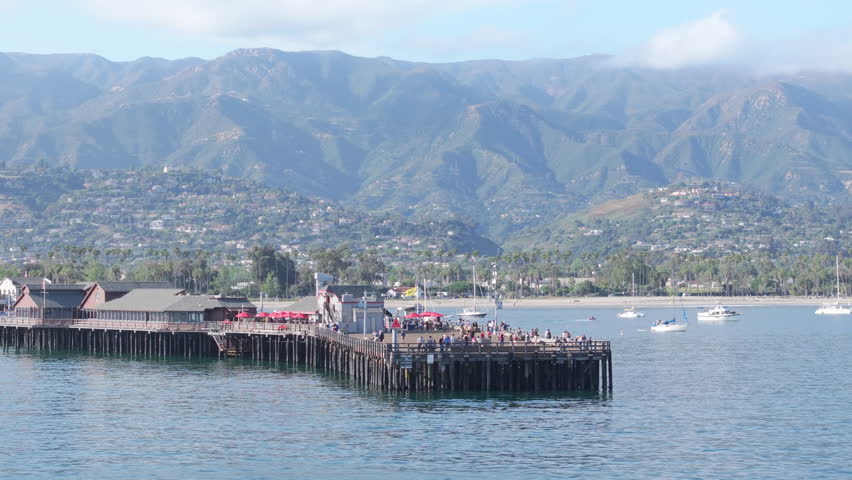 Picturesque view of Stearns Wharf with mountains, Santa Barbara, California, USA. Drone shot of old wooden pier in harbour of the city. Luxury yachts docked in Santa Barbara bay. Tourist distanation  Royalty-Free Stock Footage #1112007867