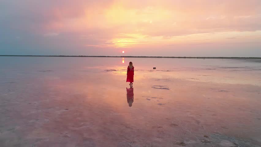 Aerial of a young woman in red dress standing in the water of a unique pink salt lake. Sunset at lake Bursol with beautiful reflections on calm water surface. Stunning scenery