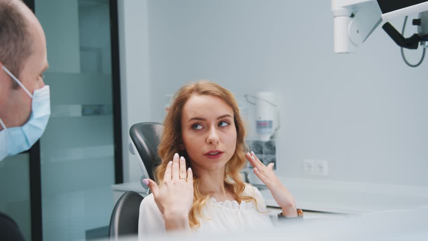 Blonde young woman complaining about toothache explaining her problems to professional dentist. Orthodontist in protective face mask listening. Stomatology. | Shutterstock HD Video #1112014927