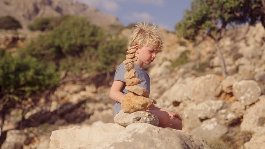 A boy is seated beside a pile of stones, witnessing them tumble down under the daytime sun | Shutterstock HD Video #1112016707