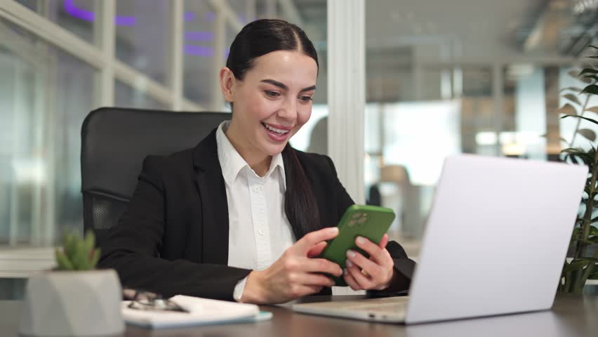 Happy female freelancer closed in black jacket sitting in front of laptop and looking at screen of modern smartphone indoor. Caucasian manager rejoicing at achieved goals in online project in office. | Shutterstock HD Video #1112017313