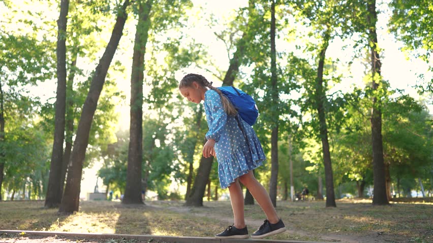 Little girl walking along the curb. concept of happy childhood and loving family. a schoolgirl in a dress and with a blue backpack walks along the lifestyle curb, keeps her balance | Shutterstock HD Video #1112020475