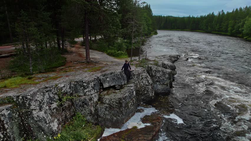 Boy sits alone on rocky bank along a road in the woods of Dalarna Sweden. Caucasian child looking down over the Storån river with swirling water. Relaxing outdoors in nature concept, aerial arc shot