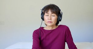 Stylish mature woman looking at camera wearing headphones using remote control changing television channels enjoying watching movie sitting in comfortable room. Attractive middle age asian female. 