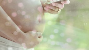 Bokeh light spots over midsection of pregnant caucasian woman using smartphone in sun. Pregnancy, self care, expectancy, communication, motherhood and healthcare digitally generated video.