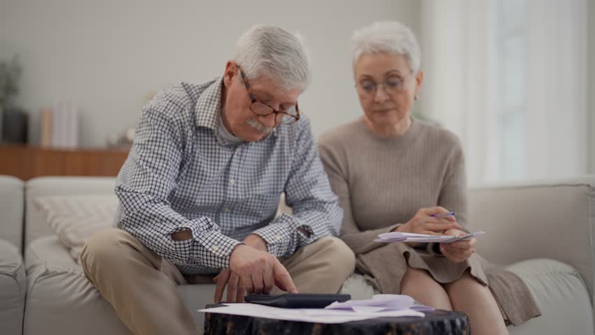 Worried senior retired couple checking calculating bills sitting on couch at home. Do paperwork discuss unpaid debt taxes, stressed family. Financial difficulties, money problem in elderly age concept | Shutterstock HD Video #1112032421