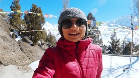 Portrait of teenager Indian girl vlogging while standing in Shashur monastery and snowy Himalayas mountains in Lahaul, India. Travel vlogger vlogging in winter wearing warm clothing and sunglasses.