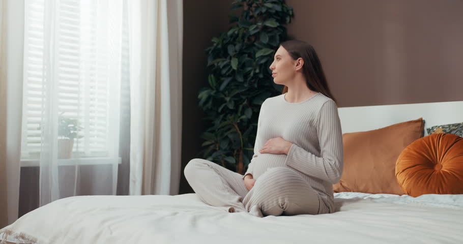 With a serene smile on her face, a pregnant woman with short hair sits on her bed, finding peace and connection with her unborn baby as she tenderly touches her belly. Concept of moment of serenity. | Shutterstock HD Video #1112036505
