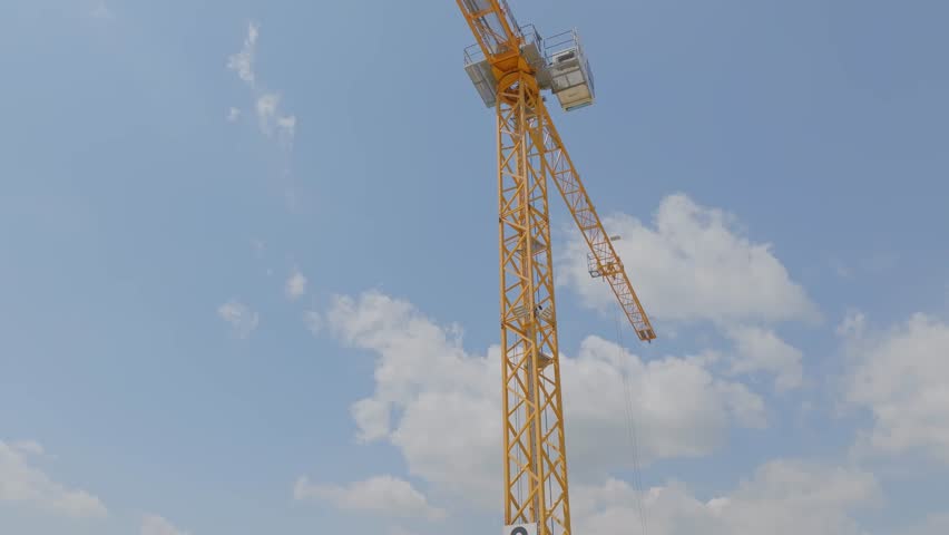 Yellow tower crane on the construction site, against blue sky with gently moving clouds. Tall yellow metal structures used for lifting and moving operating and turning.