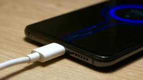USB charging cable connected and transferring power to smartphone, with quick charge blue fluid particles animation on the screen. Black color modern smartphone, lying on a wooden table while charging