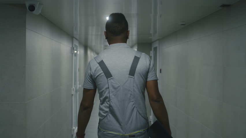 Tracking shot of African American technician in uniform walking in corridor of modern factory or plant holding digital tablet. Heavy industry worker in office building hallway at industrial facility.
