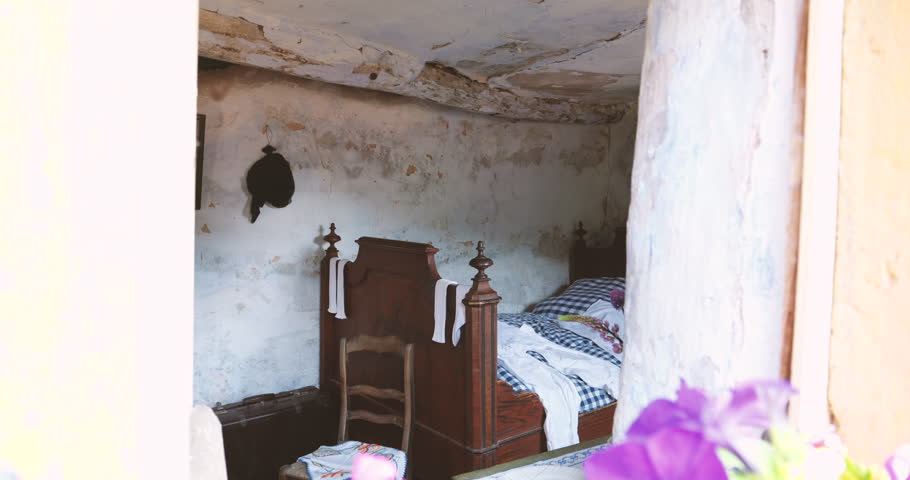 Zooming into a troglodyte house's bedroom, revealing a view through the window of an ancient bed, damaged ceiling, chair, and suitcase