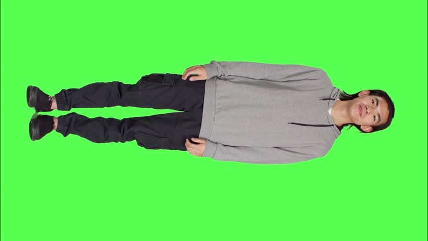 Front view of asian natural adult appears proudly on camera, smiling over greenscreen full body background. Wearing comfortable clothing, young man seems to be cheerful and peaceful with confidence.
