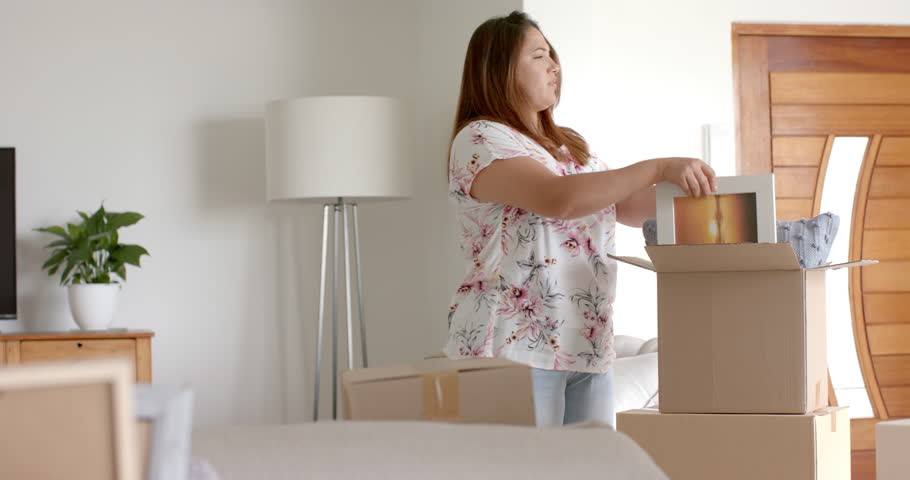 Happy plus size biracial woman unpacking picture from box in new home, slow motion. Property, moving house, new home, home ownership, domestic life and lifestyle, unaltered.