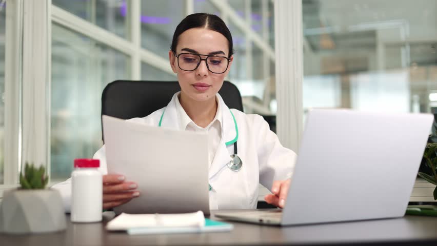 Focused woman doctor sitting at desk and working with digital device while holding documents in hand. Qualified specialist wearing stylish glasses comparing paper data with laptop in spacious room. | Shutterstock HD Video #1112053073