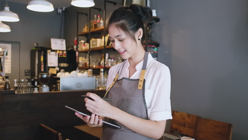 Beautiful Barista Entrepreneur Asian woman standing in cafe and using digital tablet while receive order online, looking at camera. Startup small business owner, SME business delivery online concept. Royalty-Free Stock Footage #1112053111