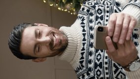 Vertical orientation of handsome Caucasian man wearing warm sweater standing near Christmas tree. Bearded male using smartphone while communicating online or browsing social media. - Powered by Shutterstock - Get 15% off with code: PIKWIZARD15