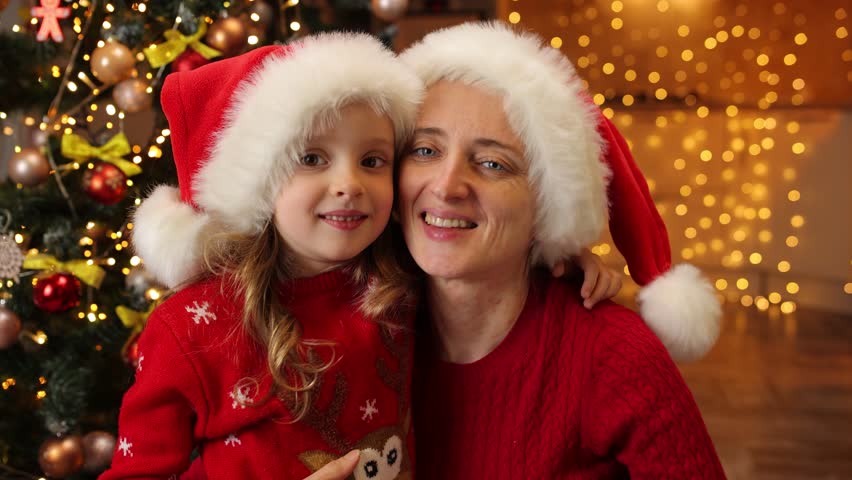 The girl is given a gift in a box and the child, surprised, thanks with kisses in return. Two girls are exchanging gifts next to a Christmas tree.