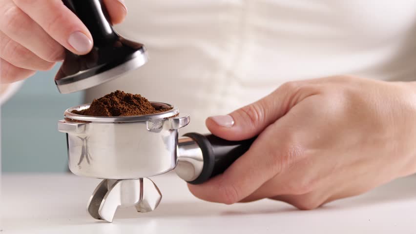 Close-up of a hand holding a portafilter with ground coffee while another hand presses it with a tamper. Making coffee at home with grinder. Enjoyment and consumption of Arabica coffee | Shutterstock HD Video #1112057561