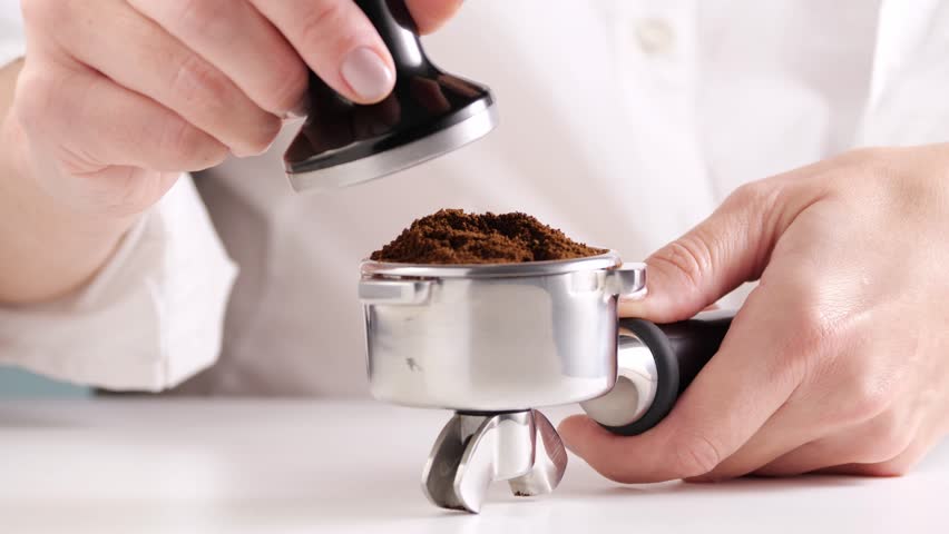 Close-up of a hand holding a portafilter with ground coffee while another hand presses it with a tamper. Making coffee at home with grinder. Enjoyment and consumption of Arabica coffee | Shutterstock HD Video #1112057575