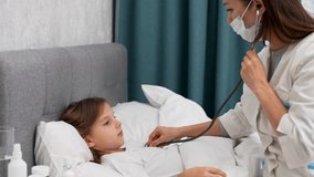 Doctor woman listening sick child girl with stethoscope sitting on bed. Pediatrician woman with stethoscope checking little patient girl at home