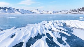 Video of scenic winter landscape of frozen Baikal lake on cold sunny day in February. Snow-capped mountains frame iced bay, and snow crusts in form of frozen waves form from wind on blue ice surface