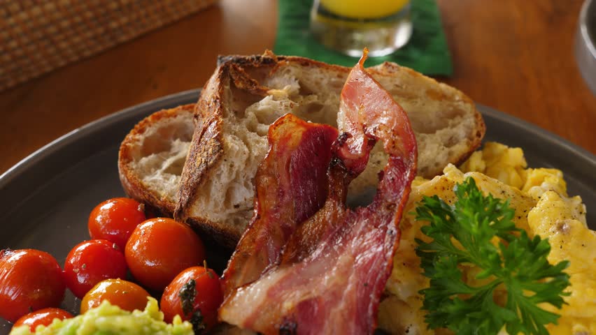 Simple and tasty breakfast, close up dolly shot of plate with pan-fried bacon, scramble egg, mashed avocado, small tomatoes and toasted slices of bread