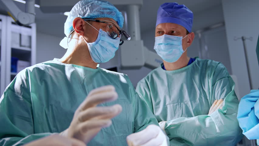 Adult surgeon wearing device glasses talks to his colleagues standing beside and listening to him carefully. Low angle view on the male doctors in surgery room.