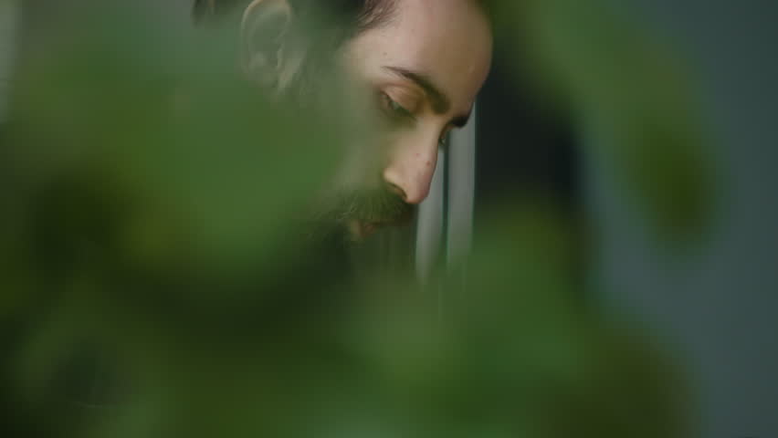 Close-up of bearded young adult man sitting in kitchen, using smart phone or writing on paper