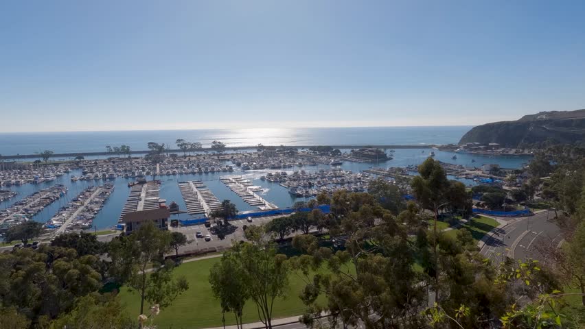 Time lapse footage of the boats and yachts sailing in the Dana Point Harbor with blue ocean water and lush green trees and cars driving at Dana Point Bluff Top Trail in Dana Point California USA Royalty-Free Stock Footage #1112066003
