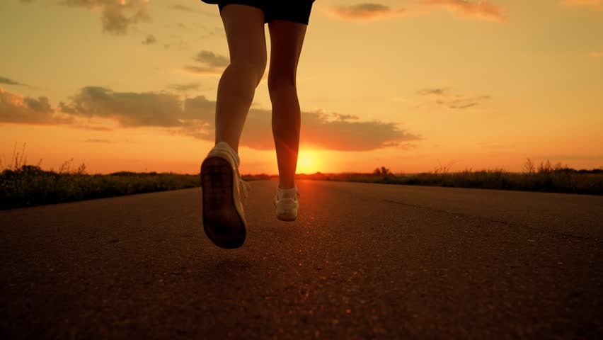 Athletes legs run along asphalt at dawn, closeup. Doing fitness, jogging on road in sun. Training jogging. Jogger girl breathes fresh air in nature. Woman runs in summer park, sunset. Jogging outside