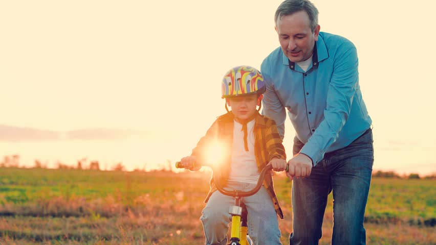 Father teaches child wearing safety helmet to ride bicycle Family in park. Child rides bicycle. Father helps his son ride bike. Boy Kid, dad play together, sunset. Child dream learns to ride bicycle | Shutterstock HD Video #1112068887