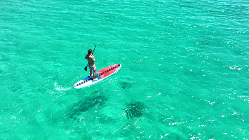 Aerial drone video of man in a paddle surf board known as Sup surfing in turquoise clear waters