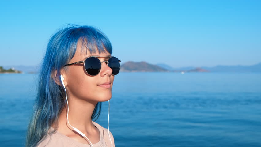 Teen with blue hair in headphones by water. A modern girl in sunglasses and earphones enjoy the free time on the beach in summer.