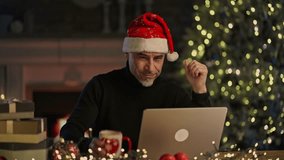 Virtual Christmas online - Happy smiling middle aged man in Santa hat at home video chatting with family on laptop computer, talking, waving hand. Cosy room with fireplace in winter. - Powered by Shutterstock - Get 15% off with code: PIKWIZARD15