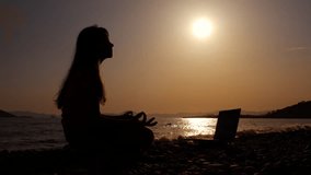 Practicing yoga against bright sunset. A teen silhouette practicing meditation with online video on laptop during nightfall time in summer.