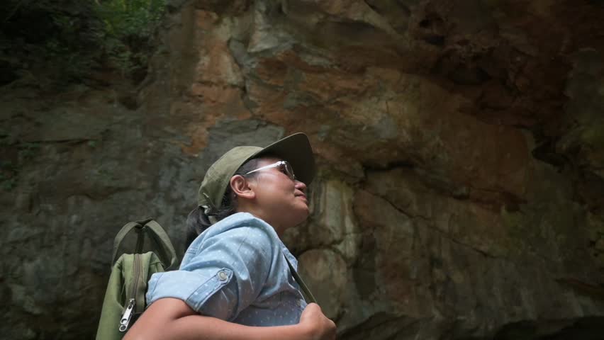 Middle aged female explorer with backpack looking around while hiking in the mountain surrounded by steep cliff during summer. Solo adventure. Outdoor pursuit. Orbit shot.