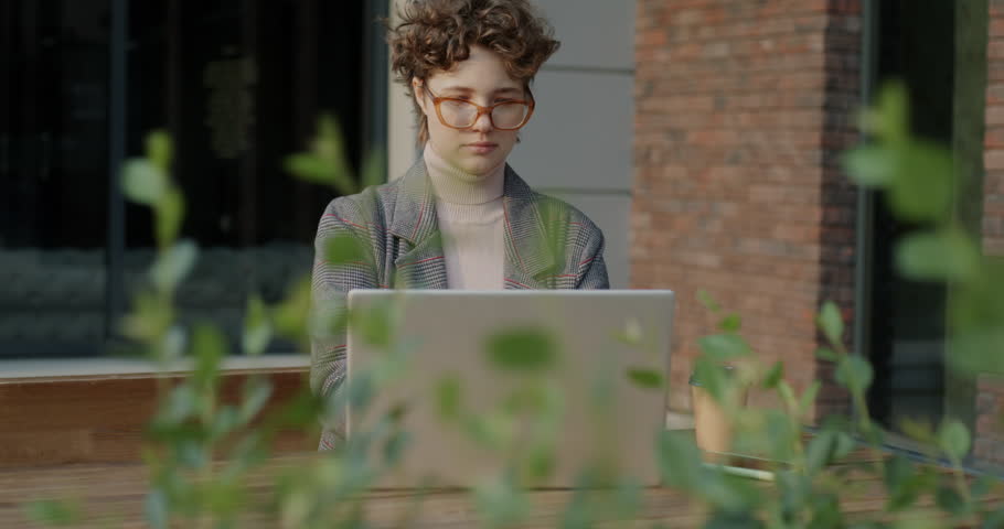 Cheerful student working with laptop computer typing and using smartphone sitting in outdoor cafe in city. Technology and online education concpt.