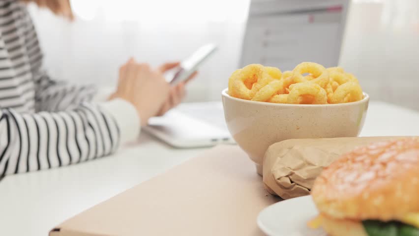 Unrecognizable woman freelancer sitting at workplace with laptop and using mobile phone during her break for dinner hamburger and onion snacks for eating at work. Royalty-Free Stock Footage #1112088055