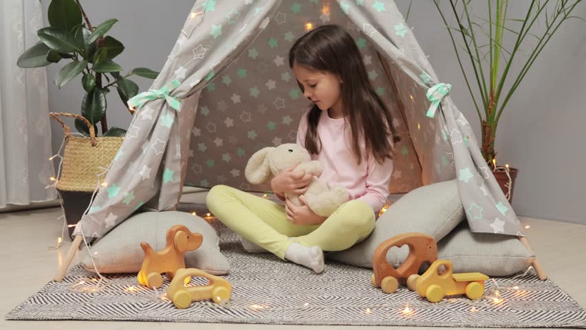 Brown haired little girl playing in teepee in living room at home shaking her soft fluffy bunny and giving him lullaby plays mother's daughter alone Royalty-Free Stock Footage #1112088081
