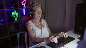 Confident, grey-haired senior woman streamer smiling while playing futuristic video game at night in gaming room, sitting indoors entertaining with modern technology