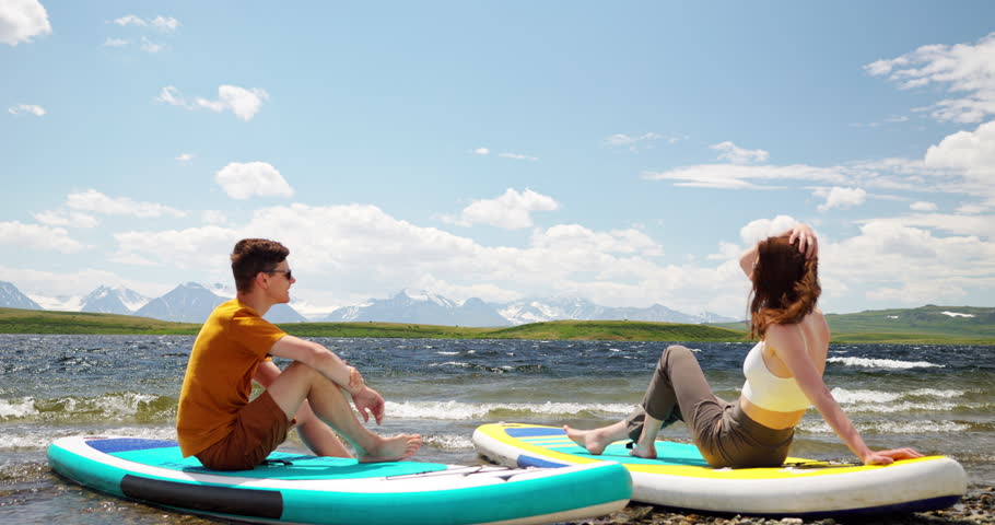 Couple of two young hikers together sit on lake shore with sup surf boards at scenic mountain landscape back view. Travel relaxation, recreation and wild water sport