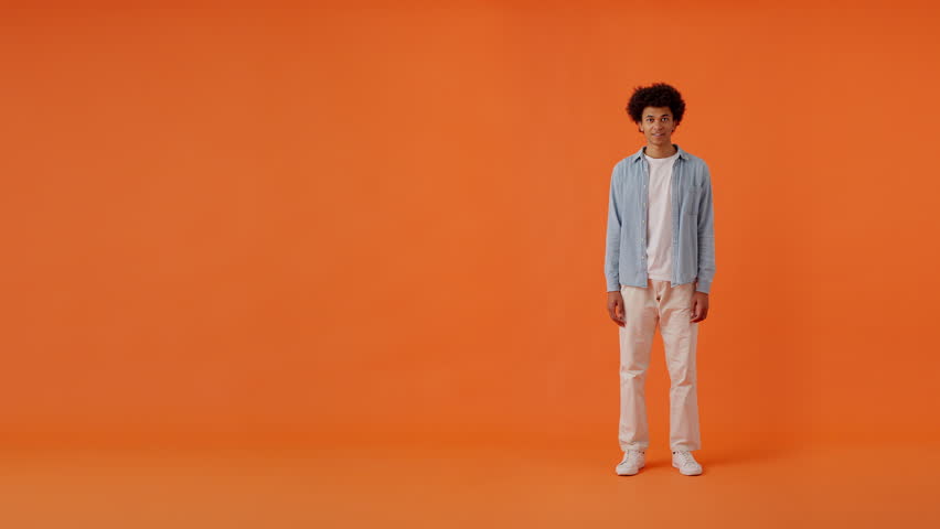 Positive multiracial man with trendy afro hairstyle in casual clothes standing in studio and pointing with finger on empty space over orange background. Concept of advertising. Royalty-Free Stock Footage #1112100845