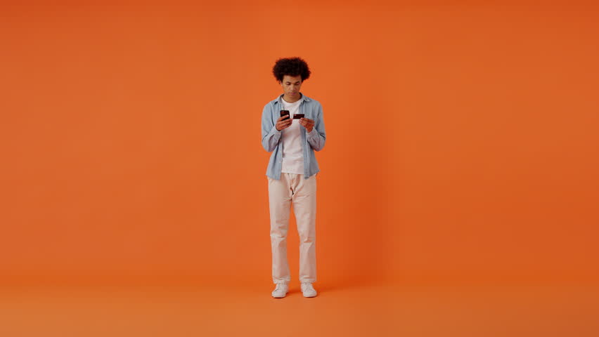 Delighted african american man using credit card and smartphone for online shopping in studio. Young satisfied male having jolly mood after paying for order in internet over orange background. Royalty-Free Stock Footage #1112100893