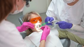 Dentist performing preventive dental care on  little girl using medical tools . Assistant helping her. Dental clinic child patient visit. Oral health care and medicare industry concept 4K video.